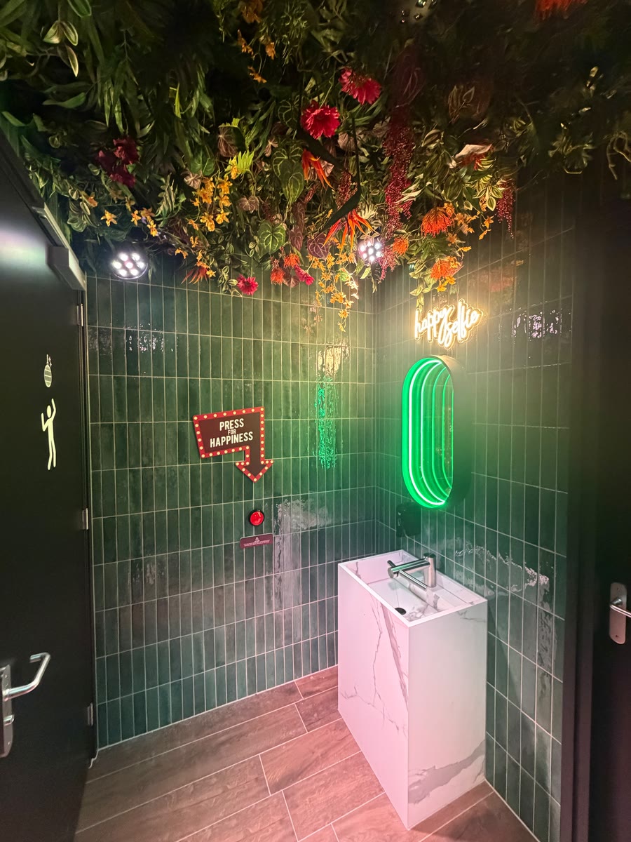 De discoWC in rust: bloemenplafond, groene tegels, de neonspiegel, het bordje Press for Happiness en de rode knop